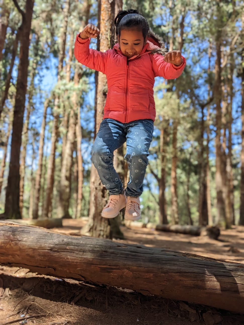 Child in a red jacket mid-jump over a fallen log in a pine forest